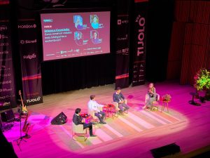 Painel 02 "Urbano e Conectado: Como construir cidades mais inteligentes e sustentáveis" no Tijolo Business 2025. No placo temos 5 pessoas sentadas lado a lado. 